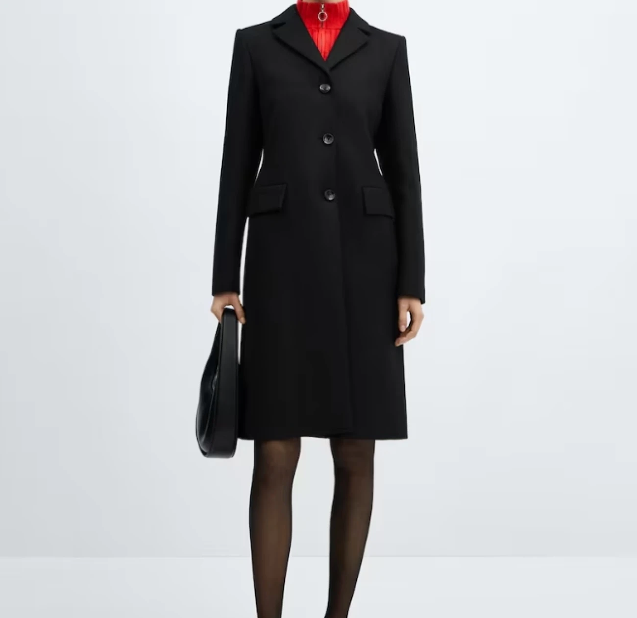 A model poses wearing a Mango black coat with a fantastic tailored silhouette and a black round bag. She has a red turtleneck