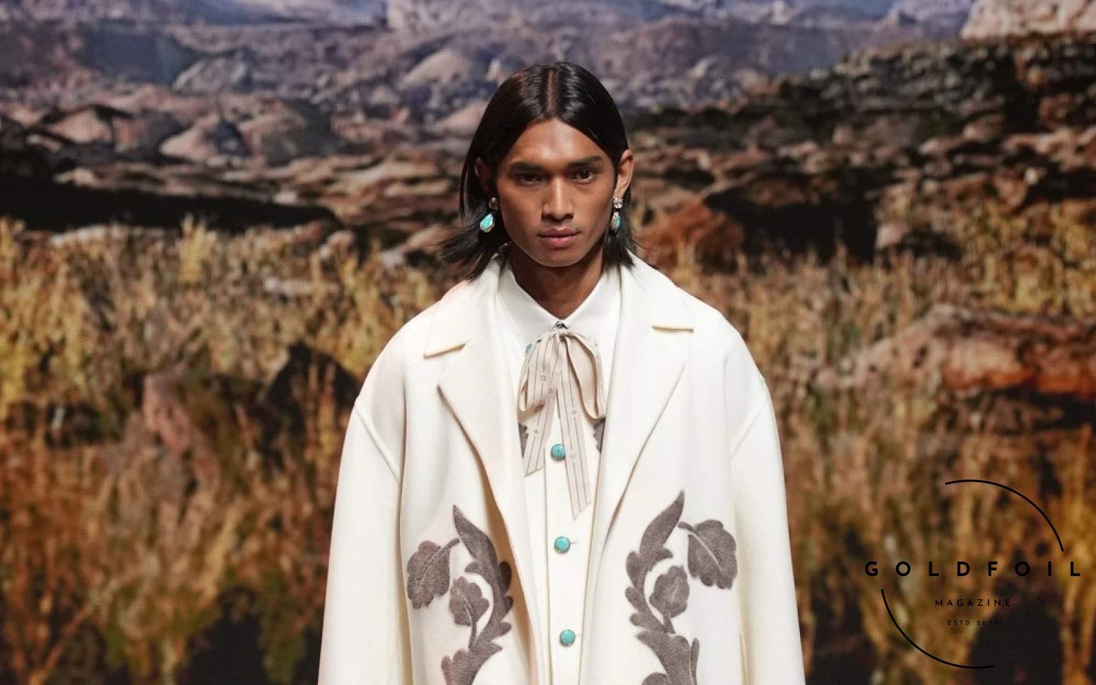 A model walks the Louis Vuitton runway show wearing a long white coat with leaves embroidery with a fringe, straight denim jeans, and a pussy bow tie shirt A model walks the Louis Vuitton runway show wearing a long white coat with leaves embroidery with a fringe, straight denim jeans, and a pussy bow tie shirt