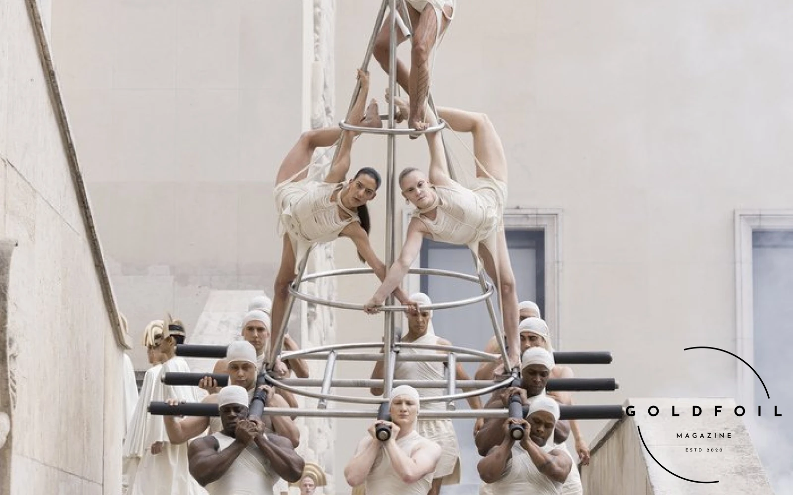 The Rick Owens 2025 Spring Summer fashion show taking place in the Palais de la Tokyo in Paris, this photo showing a human tower with wrestlers carrying models, on top a model wearing a full white outfit waving a white flag featuring a white and black hand shaking The Rick Owens 2025 Spring Summer fashion show taking place in the Palais de la Tokyo in Paris, this photo showing a human tower with wrestlers carrying models, on top a model wearing a full white outfit waving a white flag featuring a white and black hand shaking