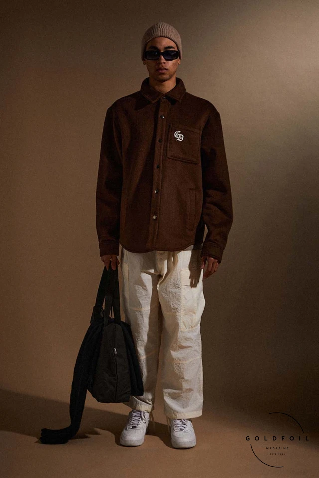 Model wears brown varsity jacket, with white cargo trousers and white trainers, with a tote bag in black and a beanie. Criminal Damage, London's iconic streetwear brand established in 1991, redefines the fashion landscape with its FW24  Model wears brown varsity jacket, with white cargo trousers and white trainers, with a tote bag in black and a beanie. Criminal Damage, London's iconic streetwear brand established in 1991, redefines the fashion landscape with its FW24