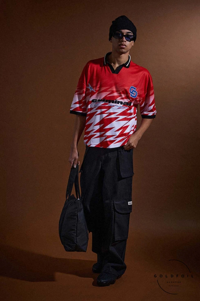 A model wears a sports polo in red and white strikes, with cargo pants and a denim tote bag. Criminal Damage, London's iconic streetwear brand established in 1991, redefines the fashion landscape with its FW24  A model wears a sports polo in red and white strikes, with cargo pants and a denim tote bag. Criminal Damage, London's iconic streetwear brand established in 1991, redefines the fashion landscape with its FW24