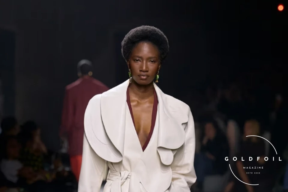 Bottega Veneta Runway show with a model wearing a white intricate trench coat paired with brown shoes and a brown leather bag Bottega Veneta Runway show with a model wearing a white intricate trench coat paired with brown shoes and a brown leather bag