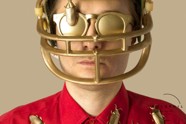 A photograph from the book Just to Land In Tokyo, featuring a character wearing a rugby helmet, gold sunglasses and covered in gold bugs