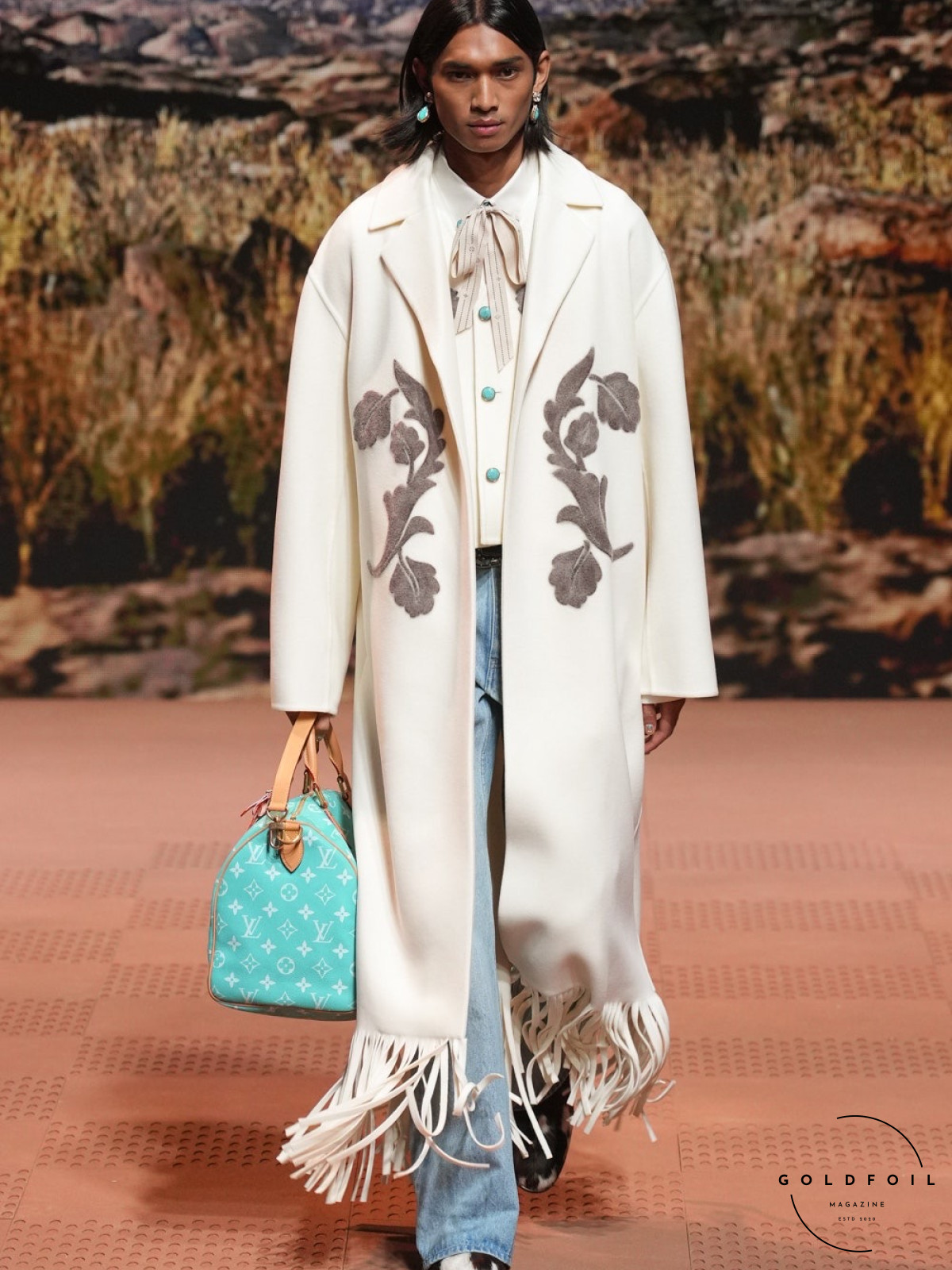 A model walks the Louis Vuitton runway show wearing a long white coat with leaves embroidery with a fringe, straight denim jeans, and a pussy bow tie shirt