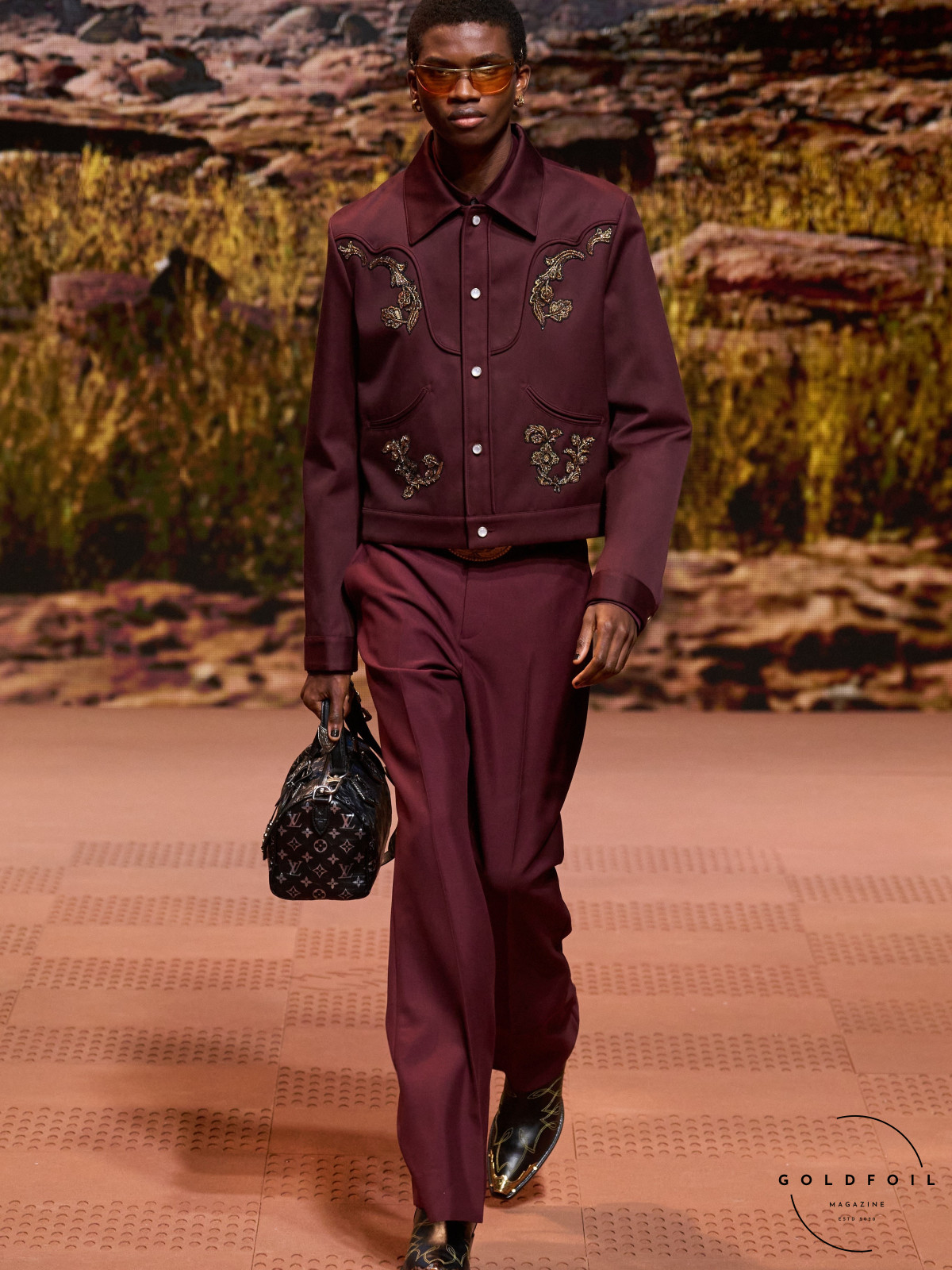 Louis Vuitton runway model wearing a red embroidered jacket and red relaxed trousers with ornamented boots and a monogram black leather bag
