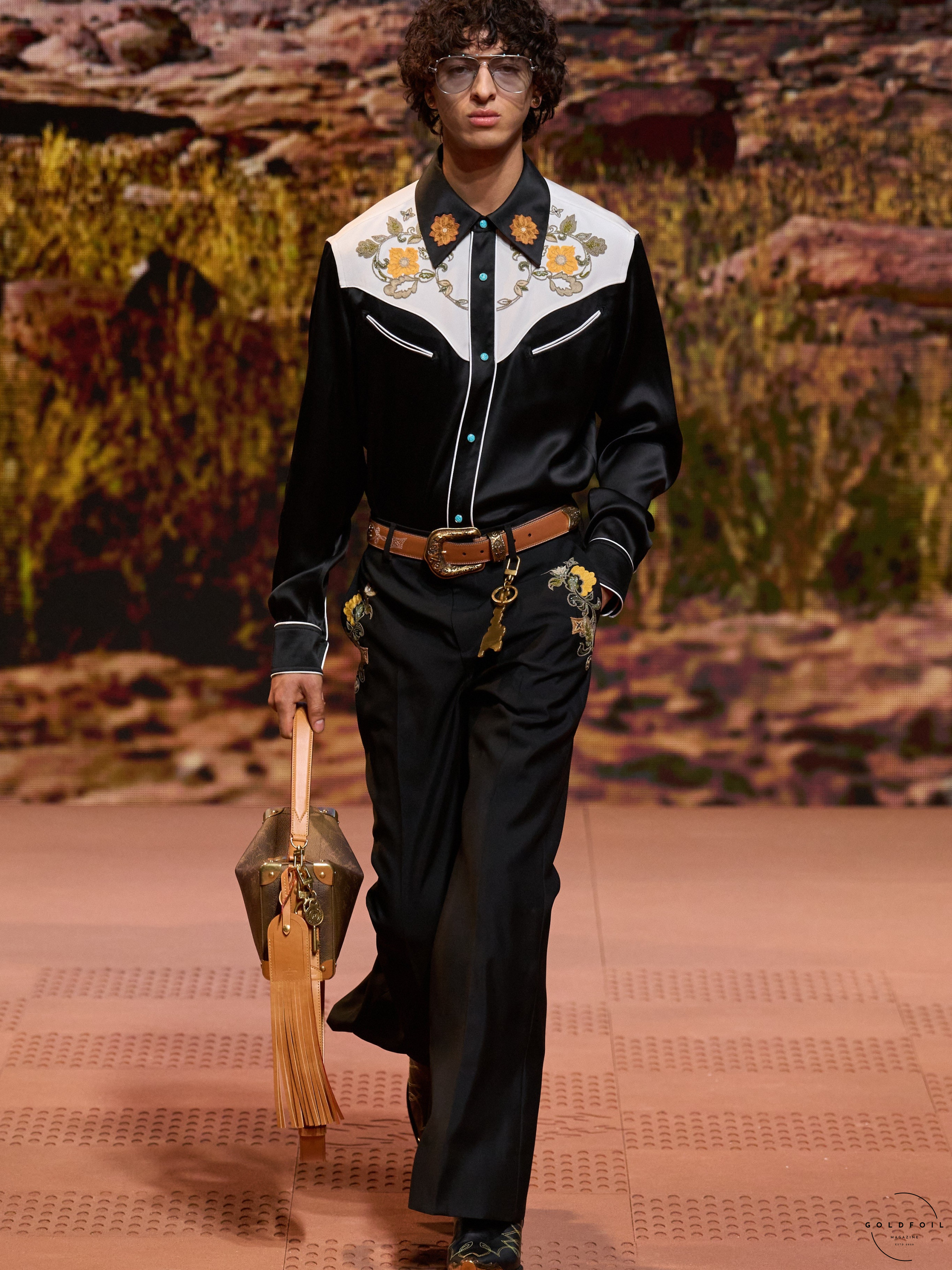 A Louis Vuitton model wearing a black and white block shirt embroidered with yellow flowers, wearing a brown leather buckle belt with a matching saddle inspired bag, and relaxed suit trousers