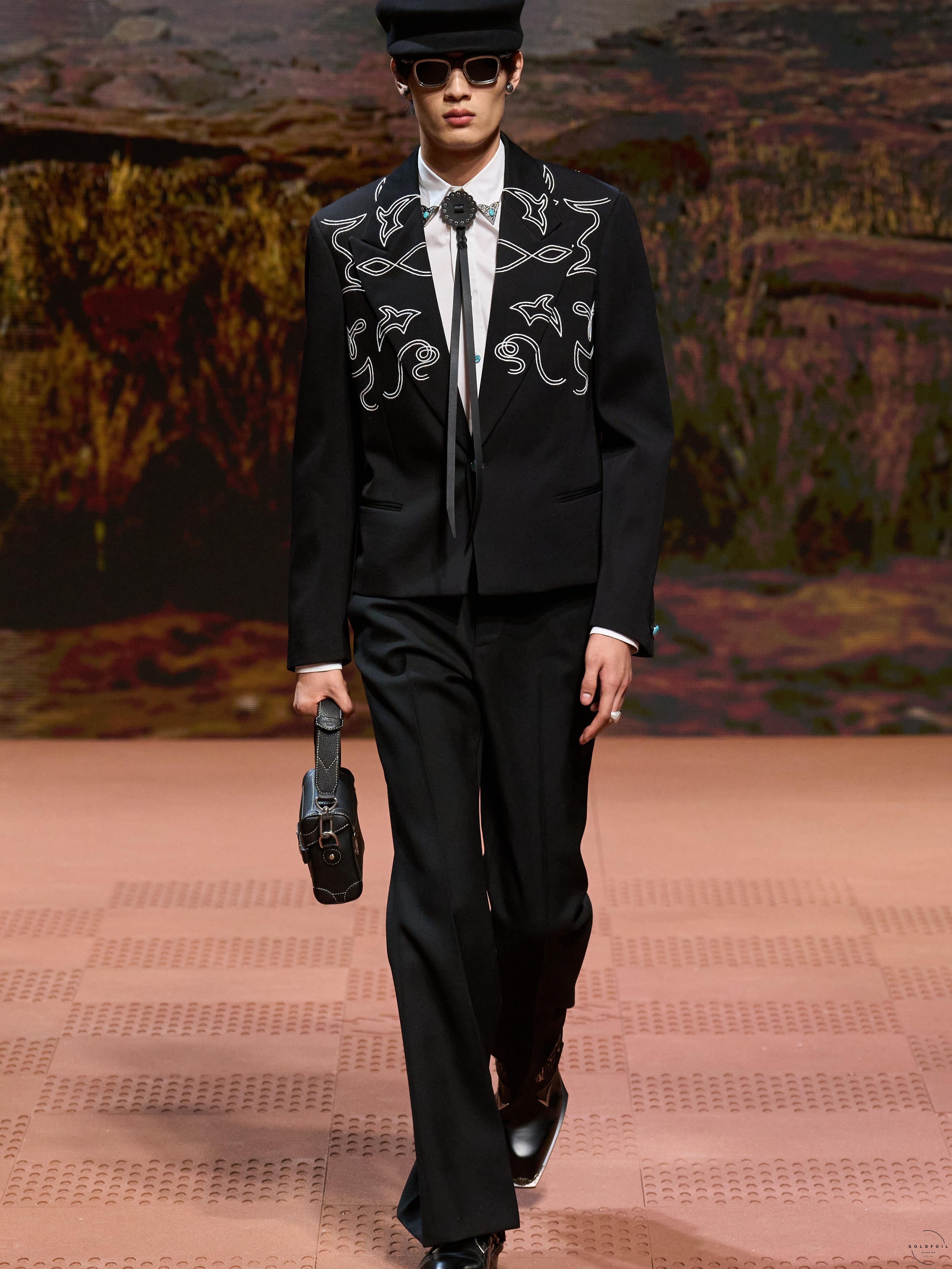 A Louis Vuitton model wearing a white shirt and a full black suit, on which the blazer is embroidered with fish like scribbles, there is a pendant clipped to the collar of the shirt with a black bow that reaches the waist