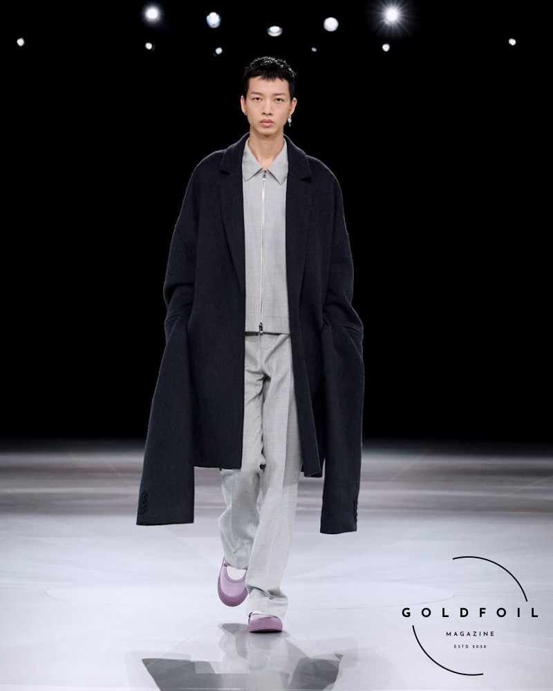 A black dior coat with long to the ground sleeves paired with a zip up collared grey shirt and straight trousers, pink shows with white socks, during Paris Fashion Week Men's Autumn Winter 2024