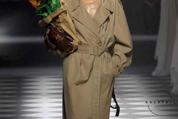 Adrian Appiolaza presented his first runway show for the Moschino brand as part of Milan Fashion Week. This is a model walking down the runway with a leather bag with green leaves and fresh bread, wearing a long trench coat