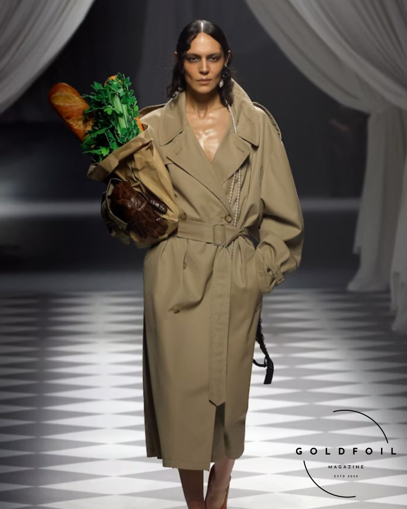 Adrian Appiolaza presented his first runway show for the Moschino brand as part of Milan Fashion Week. This is a model walking down the runway with a leather bag with green leaves and fresh bread, wearing a long trench coat